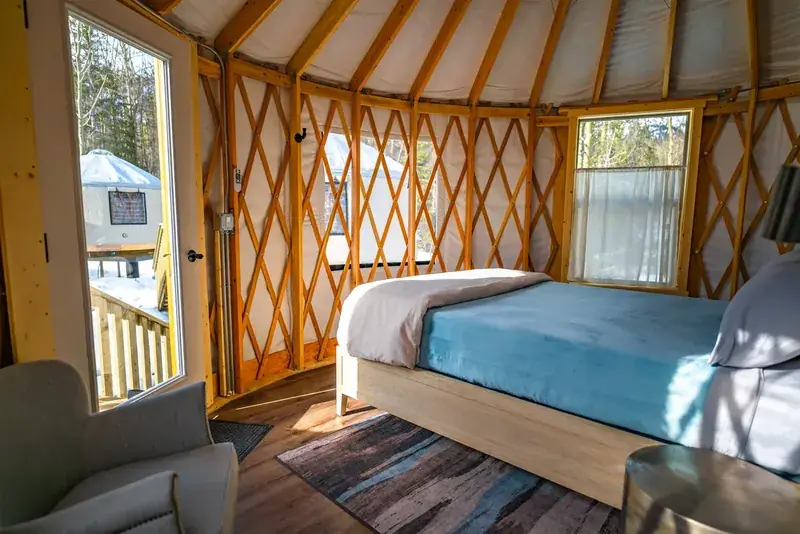 Inside a heated yurt at Alaska Rendezvous Lodge, with a made Tempur-Pedic queen bed, an armchair and a lattice timber frame, and a glass door onto a snow-covered deck facing a second yurt in the birch forest